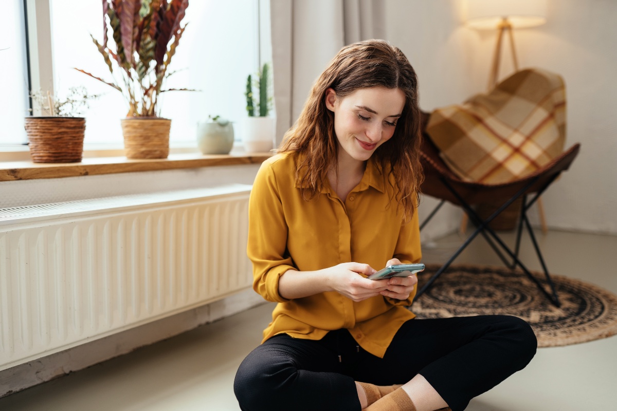 Junge-Frau-sitzt-bei-der-Heizung-im-Wohnzimmer-schaut-auf-Handy